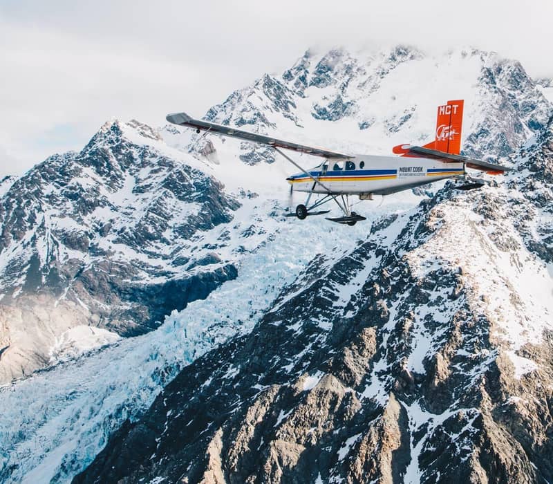 Small airplane flying near snow-covered mountains and glacier under cloudy sky