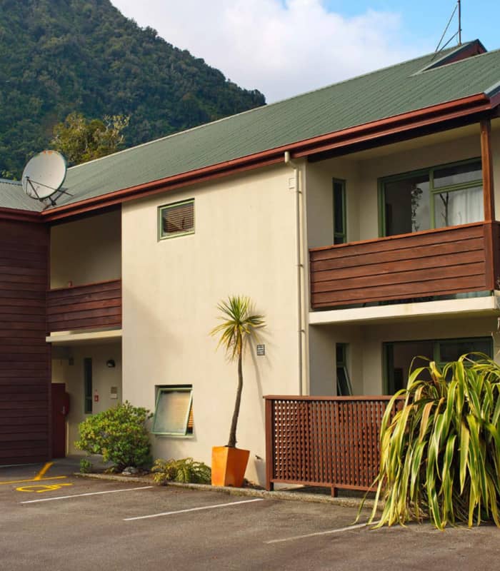 Two-story motel building with balconies and parking in front, with a green mountain behind.
