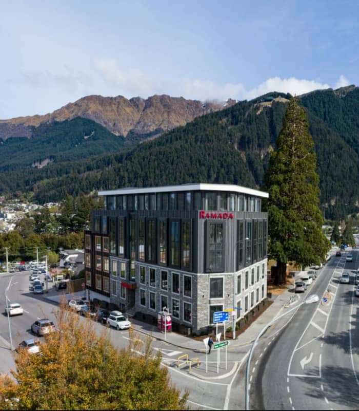 Ramada Hotel & Suites building at a street intersection with mountains and trees in the background.