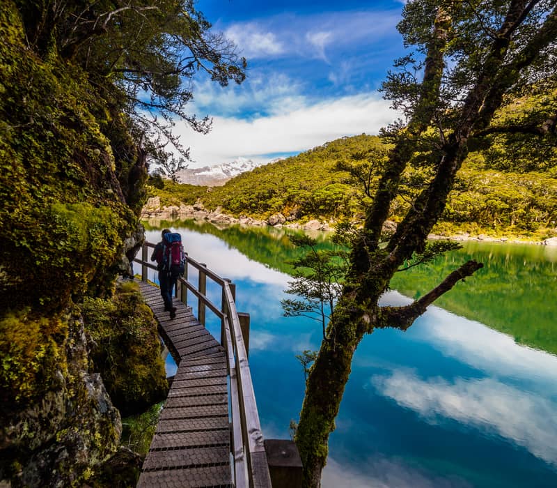 Person hiking on wooden boardwalk next to a clear lake with trees and mountains in the background
