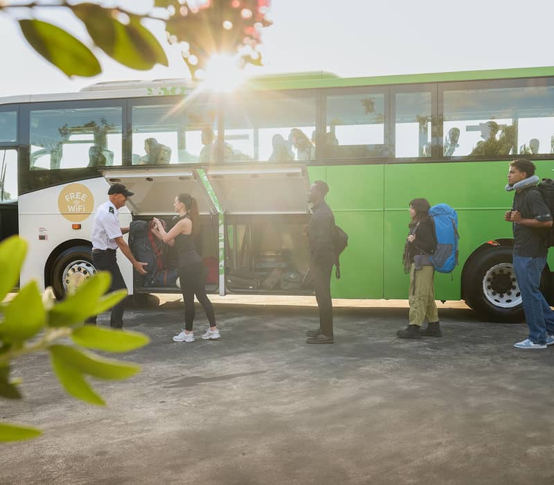 Passengers queuing to board a green Intercity bus with backpacks and luggage on a sunny day.