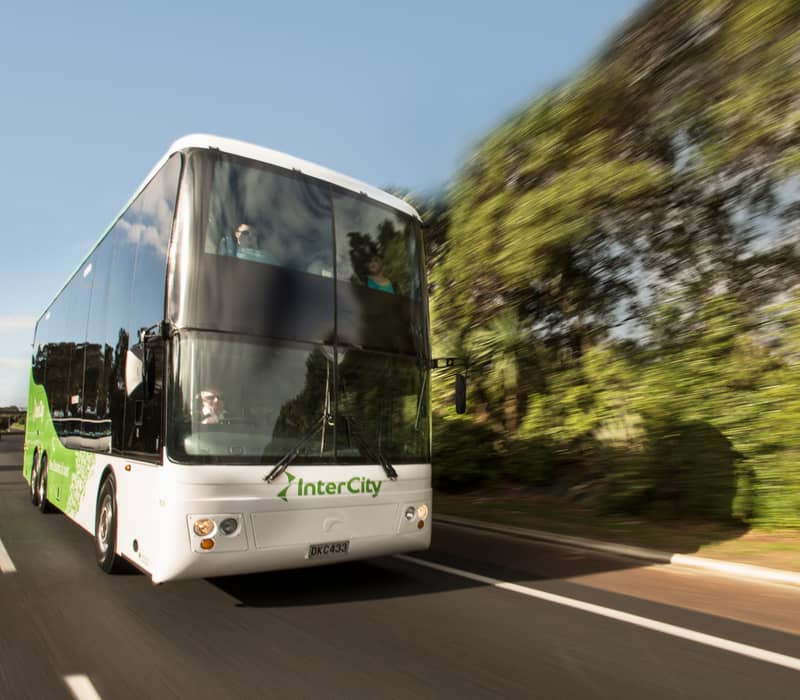 InterCity double-decker bus moving on a road with trees alongside under a clear blue sky.