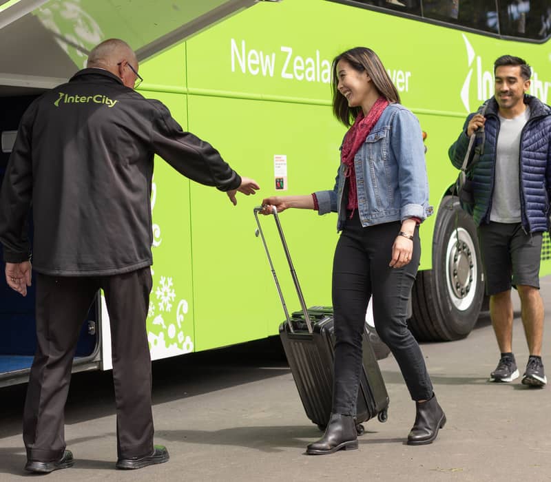 Bus driver helping woman with suitcase near green bus, man with backpack follows behind.