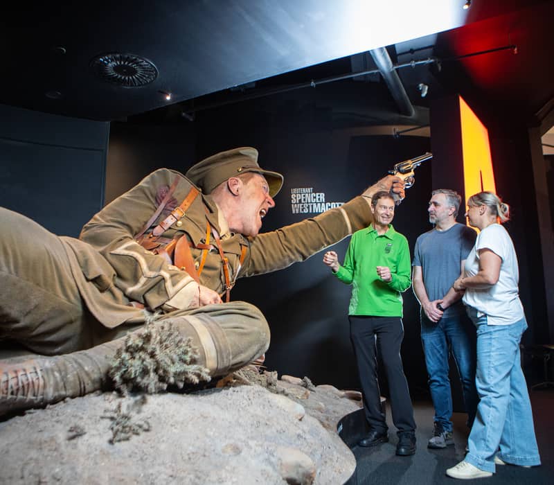Three people listen to a guide next to a large soldier statue holding a gun at a museum exhibit.