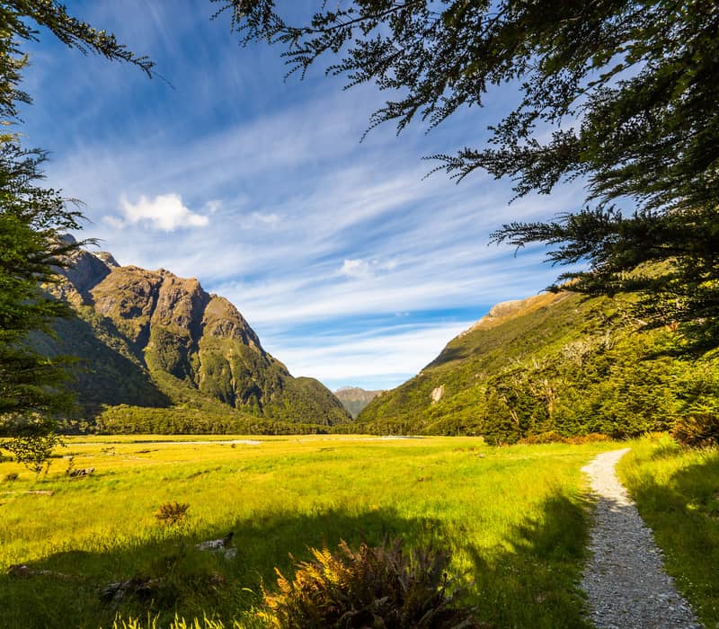 Gravel path winding through a green valley with mountains and blue sky in the background