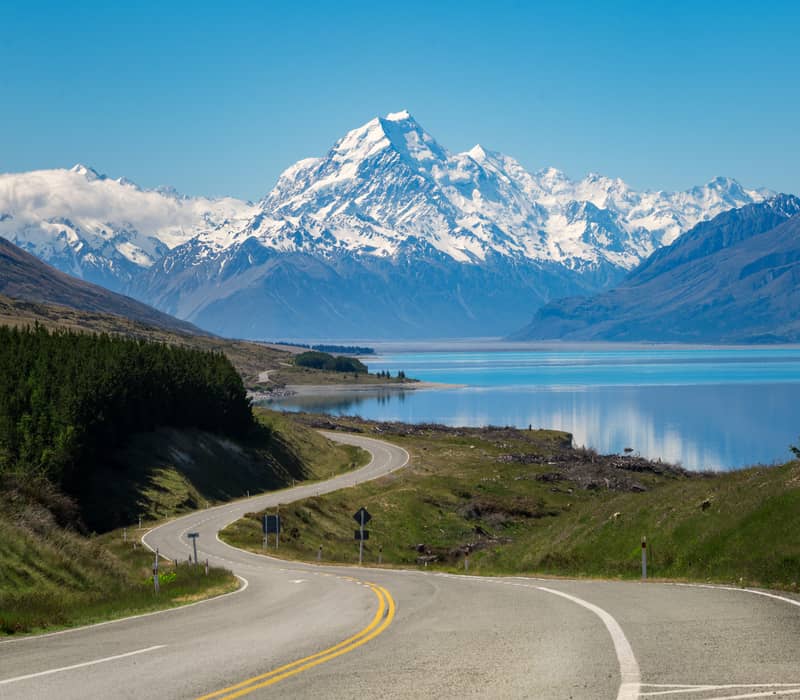 Curved road leading towards snow-covered Aoraki/Mt. Cook mountains by a reflective lake.