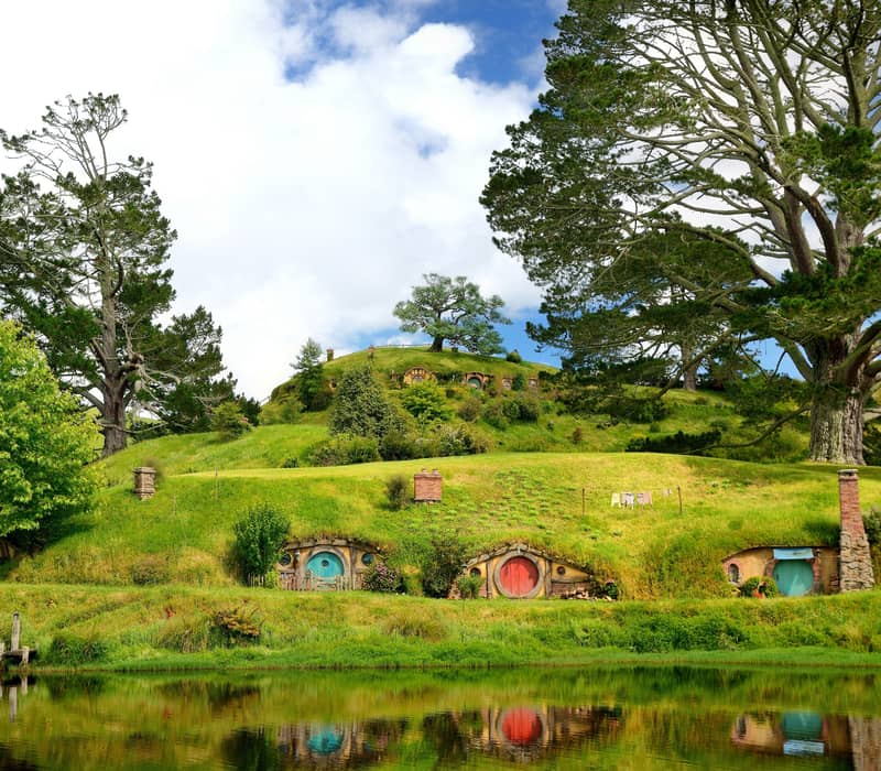 Colorful hobbit house doors built into green hills with large trees and a pond reflecting the scene