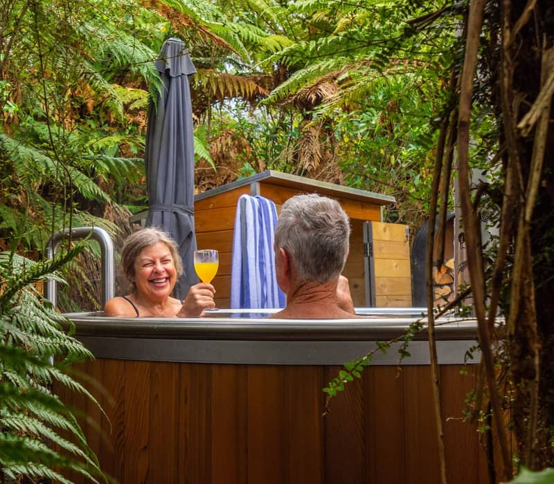Couple relaxing in hot tub in forest, one holding a drink.