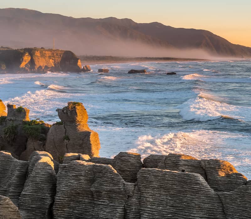 Pancake Rocks coastal rock formations at sunset with waves and mountains in the background