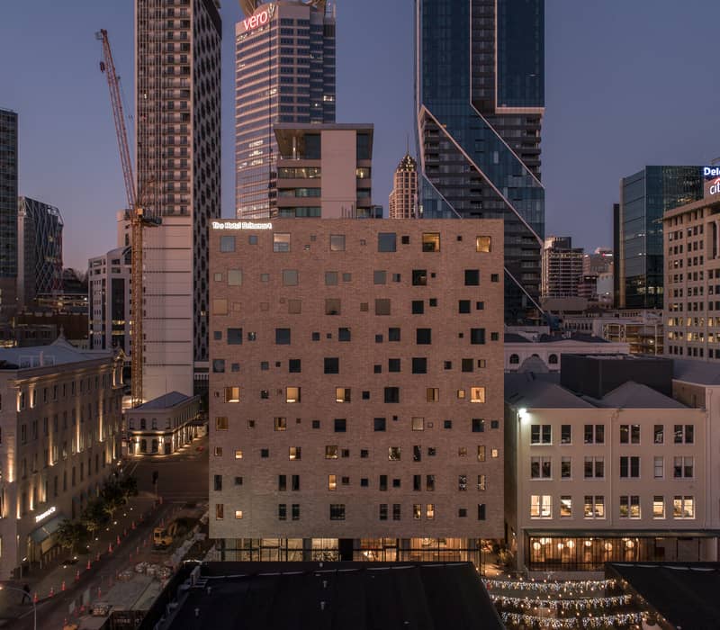 The Hotel Britomart building lit up at dusk in a city with tall buildings and cranes.