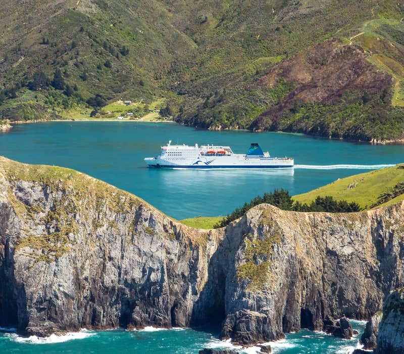 Ferry named Kaitaki sailing through a bay with cliffs and green hills in the background.