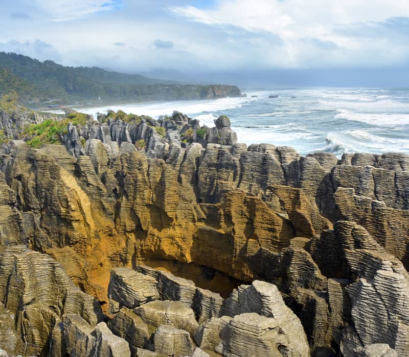 Layered limestone rock formations known as Pancake Rocks with ocean waves and green hills in the background