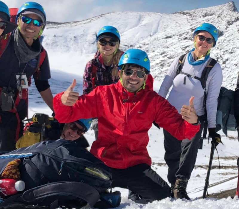 A smiling group of seven hikers in blue helmets and sunglasses posing on a snowy mountainside with rocky peaks in the background.
