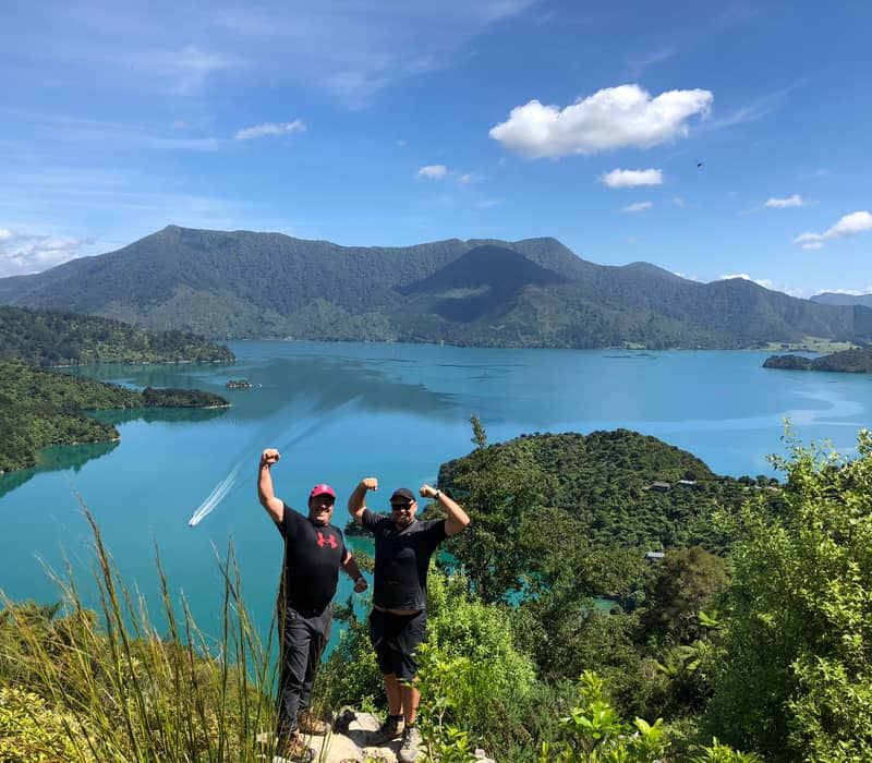 Two men flexing their arms on a hilltop with a blue lake and forested mountains behind them.