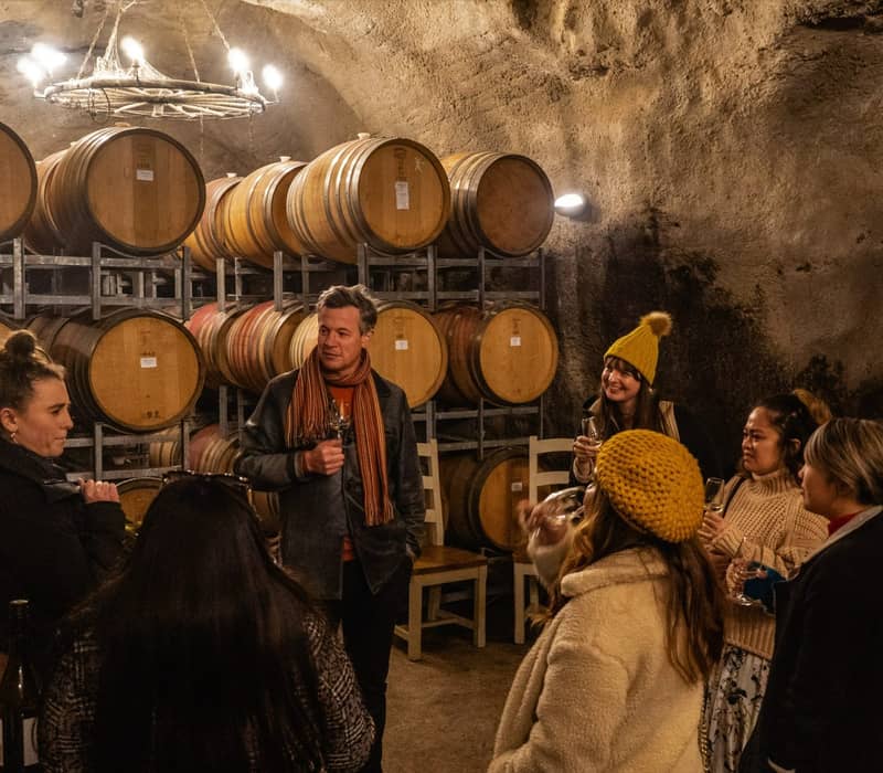 Group of people tasting wine in a cellar with wooden barrels around. Credit: Appellation Wine Tours
