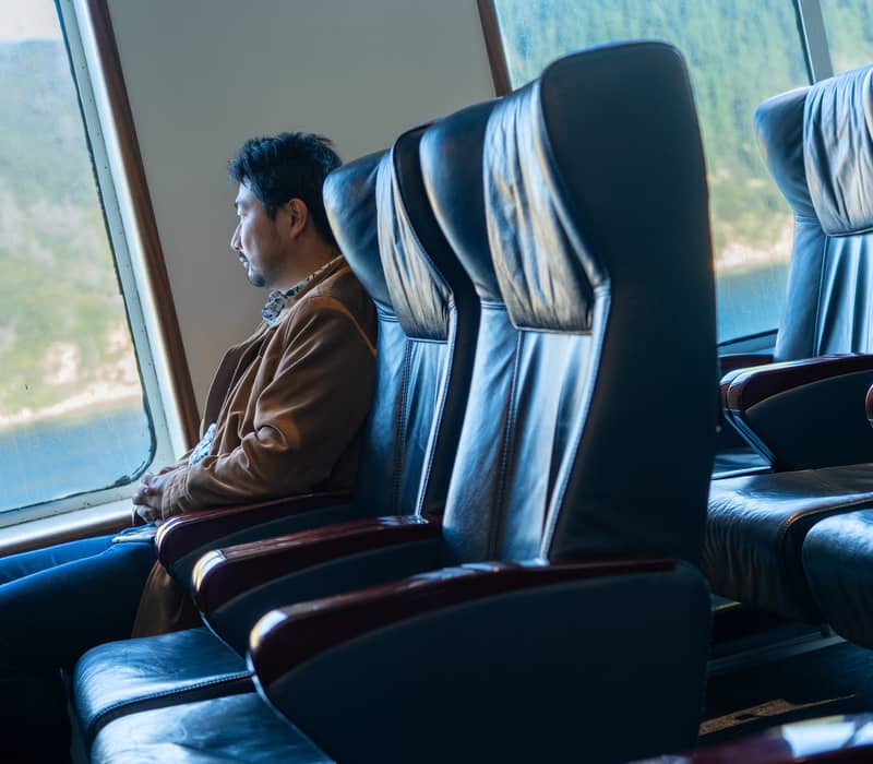 Man sitting in a boat lounge looking out a window at forest and water scenery.