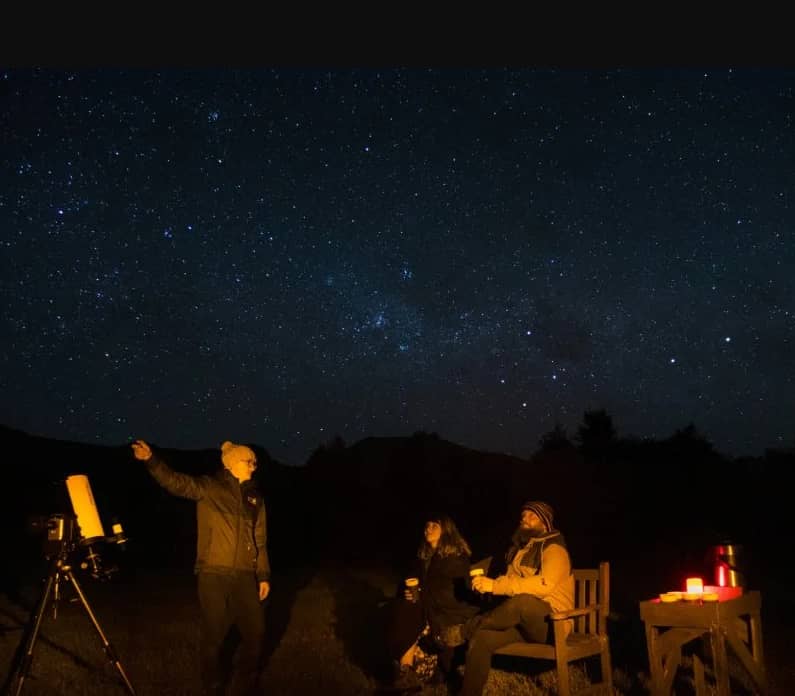 People stargazing outdoors at night with a telescope under a star-filled sky.