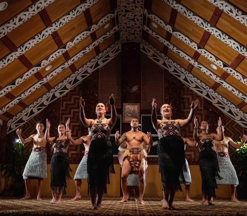 Performers in traditional Maori dress doing a group dance inside a decorated meeting house.