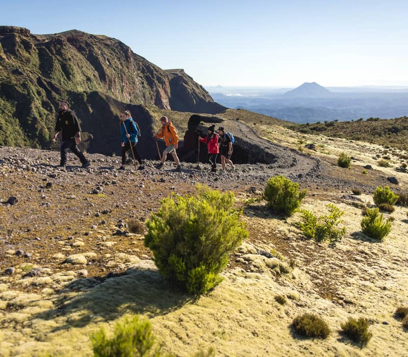 Group of hikers walking on a mountain trail in a volcanic landscape with bushes and distant hills.