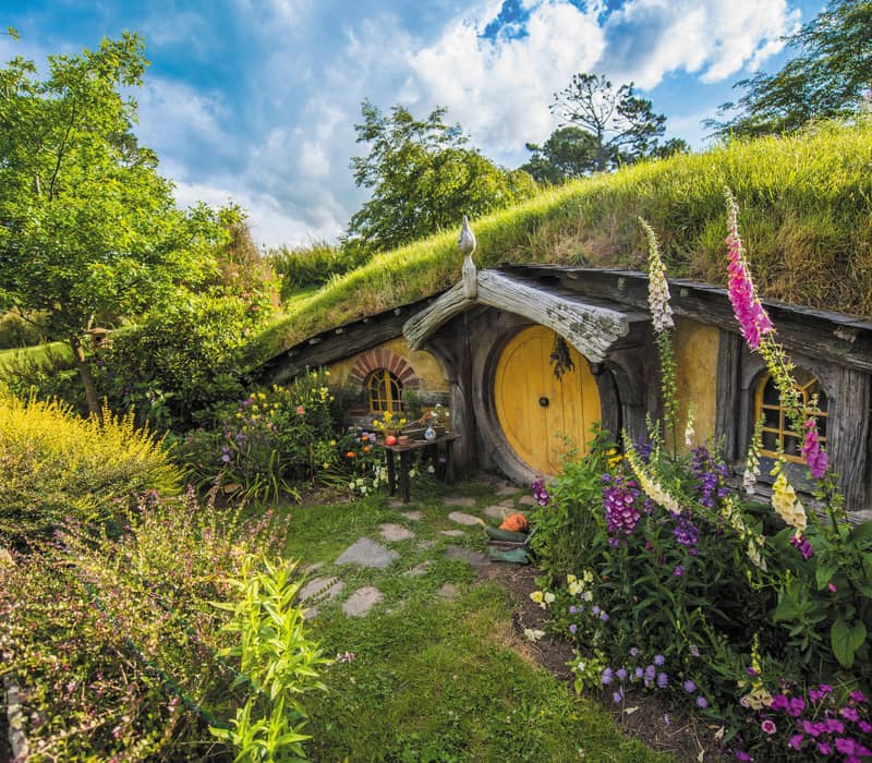 Hobbiton hobbit house entrance with round yellow door surrounded by colorful flowers and greenery under a blue sky.