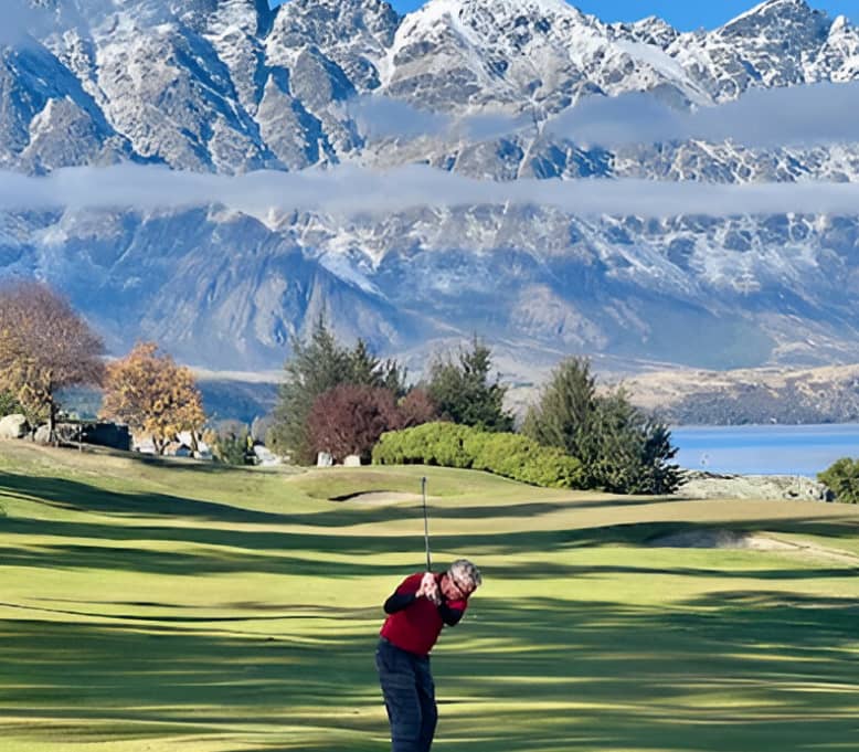 Golfer teeing off on green course with snow-covered mountains and lake behind