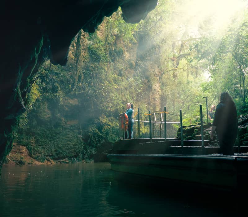 A view from inside the dark mouth of a cave looking out at an exit platform and jetty, where dramatic sunbeams slice through a lush, misty green forest canopy.