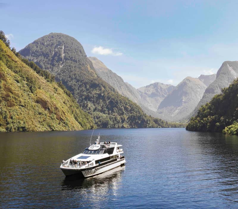 Boat sailing through calm fjord waters surrounded by green mountains under a clear sky.
