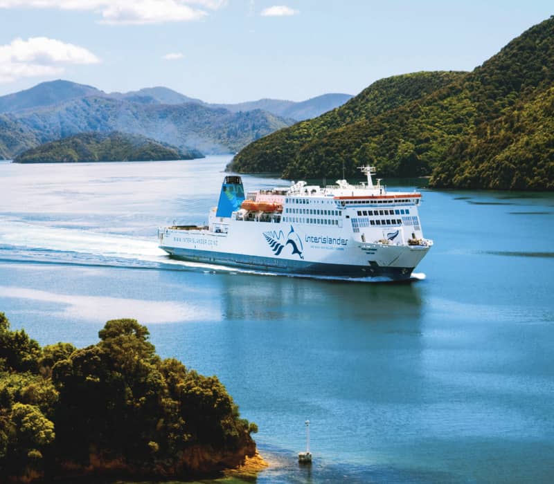 Ferry cruising through blue waters with green forested islands and mountains in the background. Credit: Interislander
