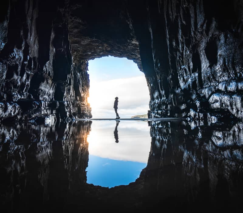 Silhouette of a person standing at the entrance of a cave with water reflection and blue sky outside.