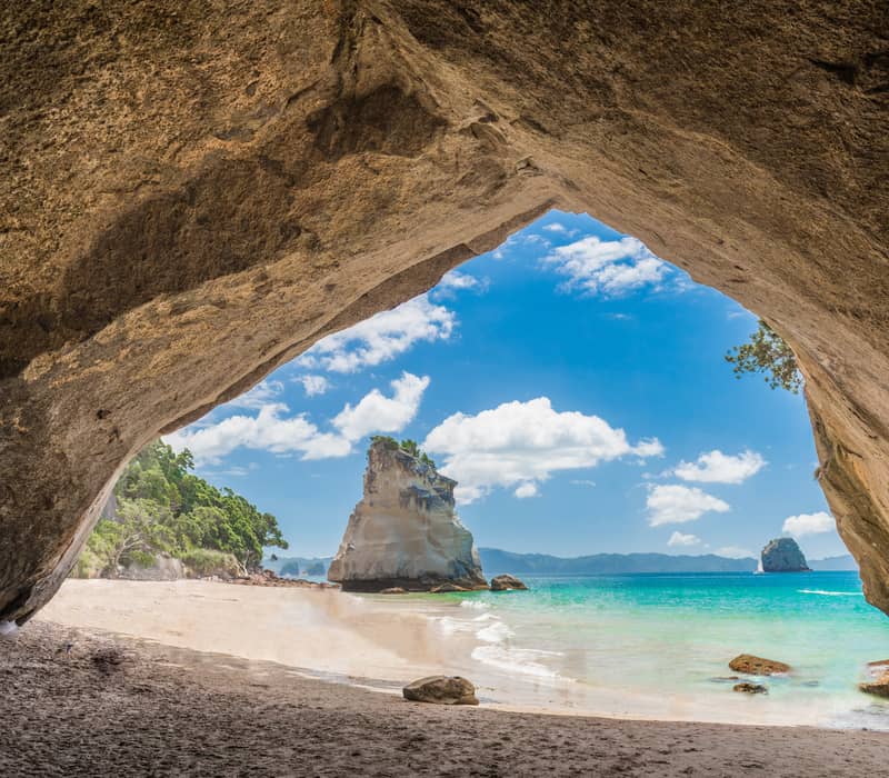 View of sandy beach and ocean seen through a large natural rock arch with sea stacks and blue sky