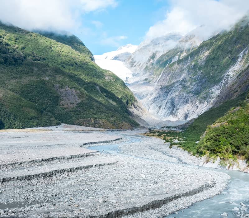 Franz Josef Glacier flowing through mountain valley with river and green forest under blue sky