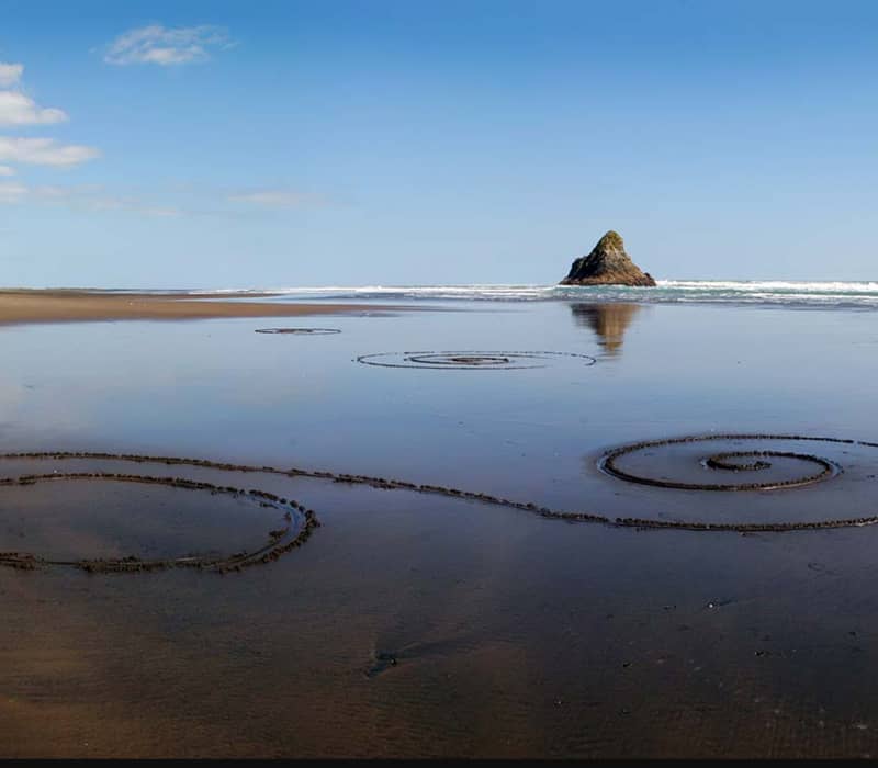 Spiral shapes drawn in the sand on a beach with a rock in the ocean and clear blue sky.