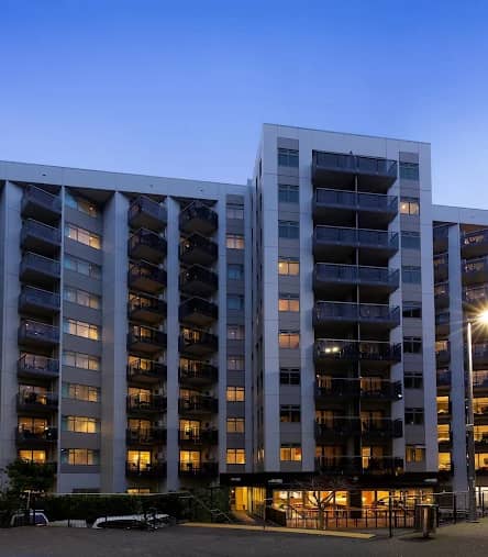 Modern hotel building with balconies lit up in the evening under a clear blue sky.