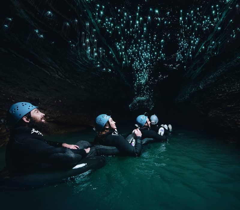 Group of people in wetsuits and helmets floating in water inside a dark cave with glowing blue glowworms on the ceiling.