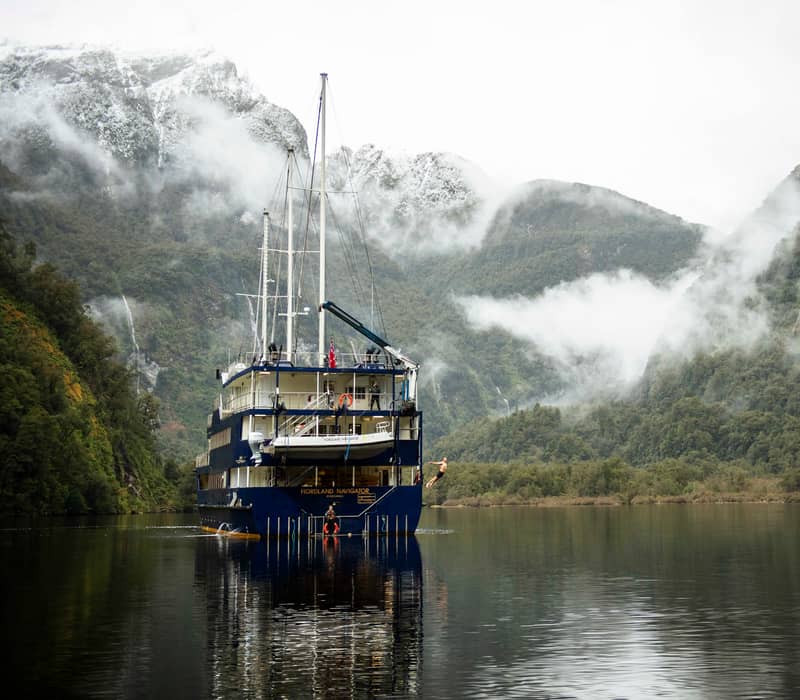 Large ship anchored in misty fjord with mountains and forests surrounding calm water.