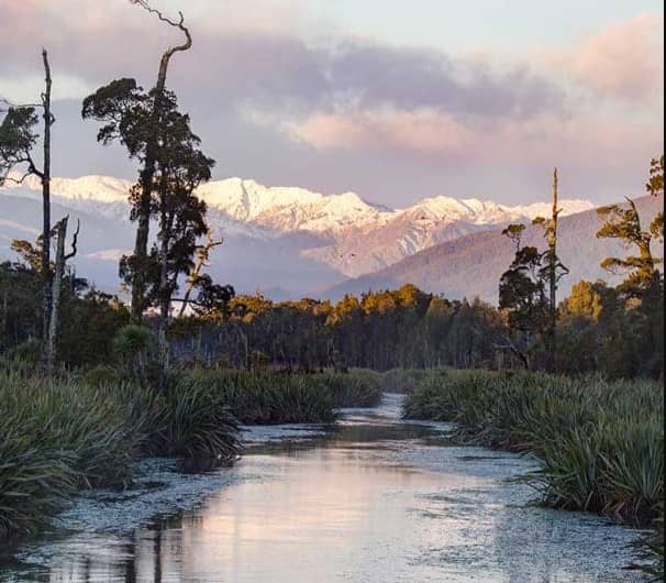 River flowing through forest towards snow-capped mountains under a colorful sky.