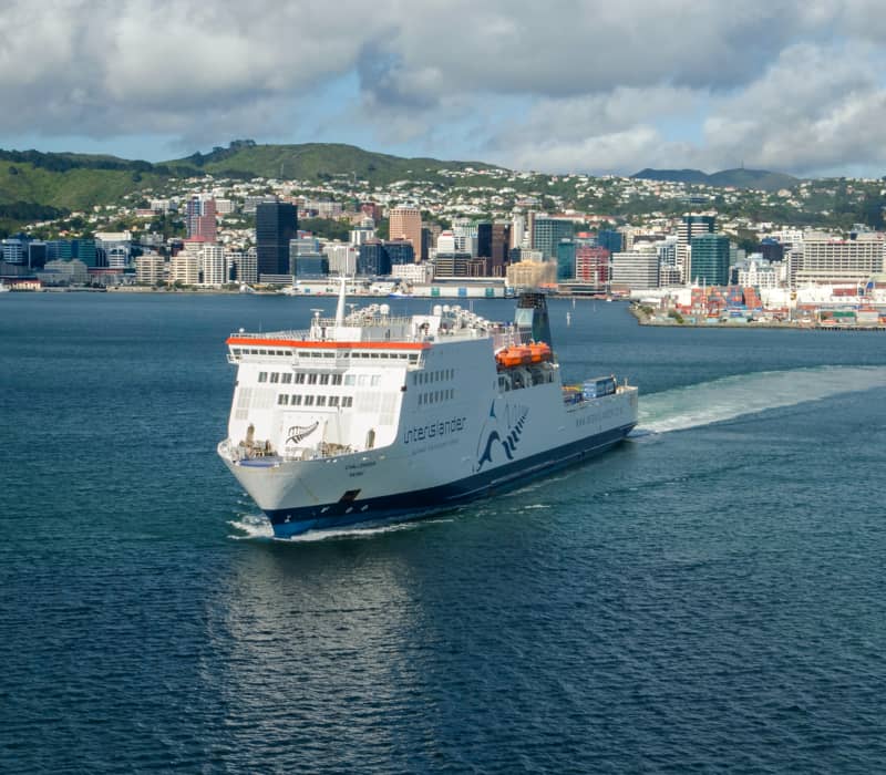 Ferry sailing in harbor with city and hills in the background under a cloudy sky.