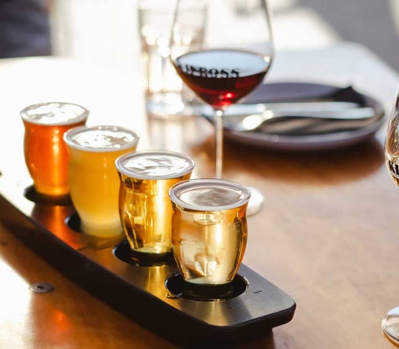 Four small glasses of beer on a wooden paddle with two wine glasses in the background on a table.