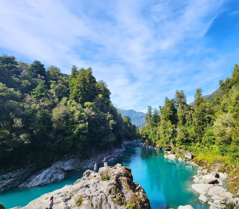 Bright blue river flowing through green forest with rocky banks and mountains under a clear sky.