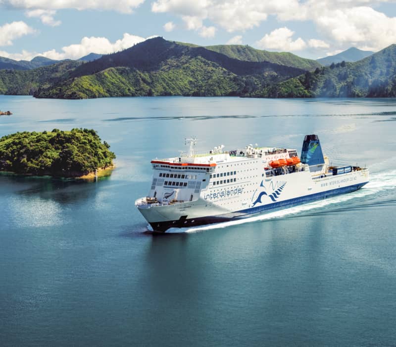 A large ferry sails through blue waters with green islands and mountains in the background under a partly cloudy sky.