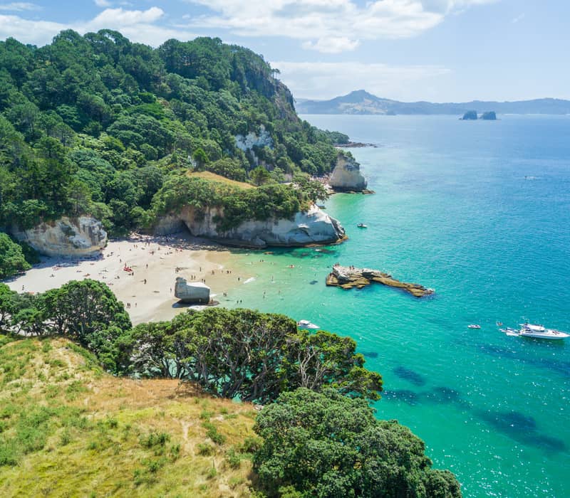Aerial view of a sandy beach with turquoise water and boats near forested cliffs at Cathedral Cove.