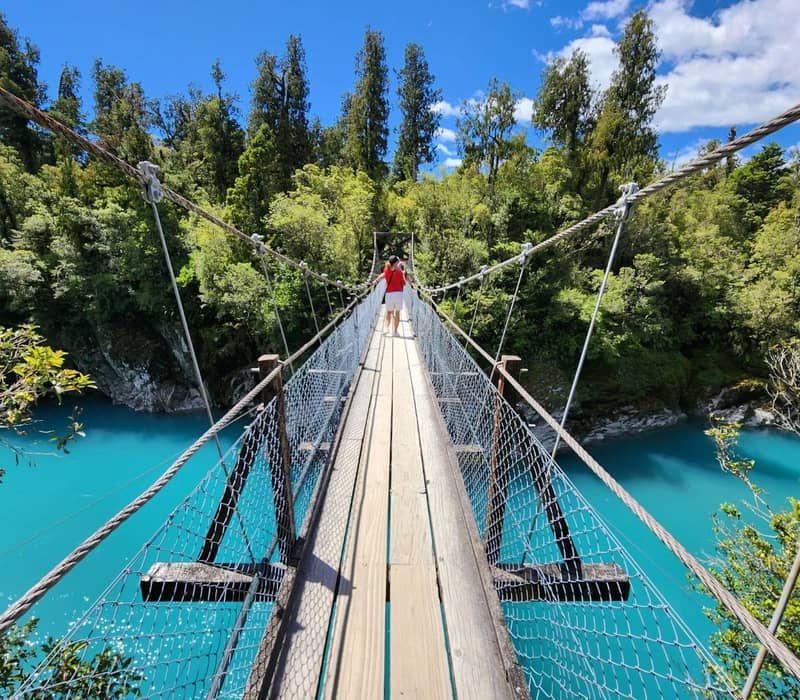 Person walking on wooden swing bridge over bright blue river with green trees and sunny sky