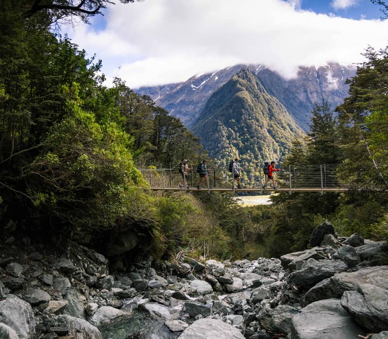 Four hikers crossing a suspension bridge over rocky riverbed with mountains and forest behind