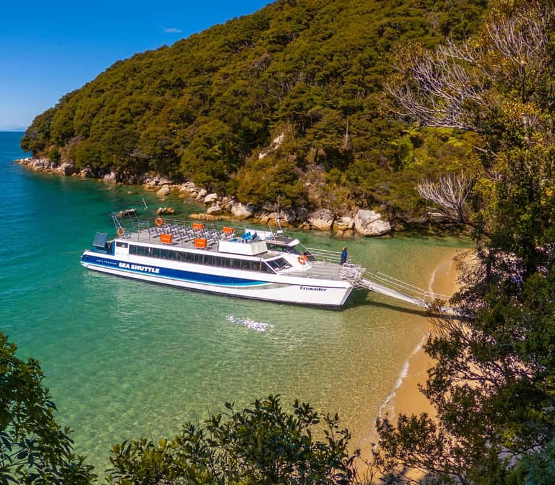 Sea shuttle docking at a sandy beach with clear turquoise water and dense green forest under a blue sky.