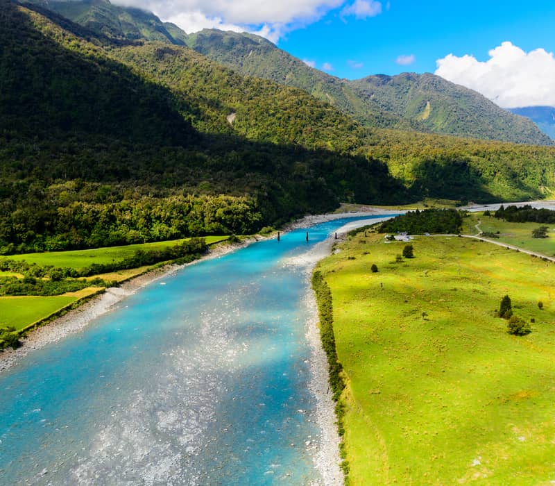 Blue river flowing through green valley with forested mountains in background under a partly cloudy sky.