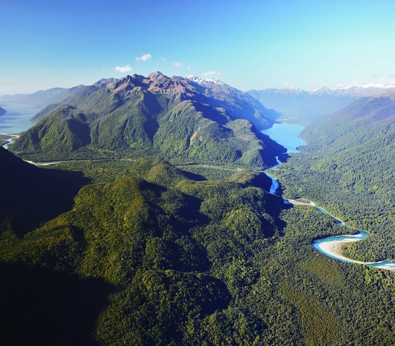 Aerial view of a valley with a winding river, forest, lakes, and mountains under a clear blue sky.