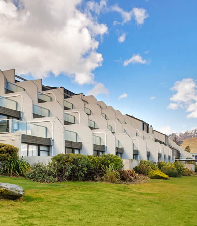 Sign for Copthorne Hotel & Apartments with snow-capped mountains and road under a blue sky with clouds.