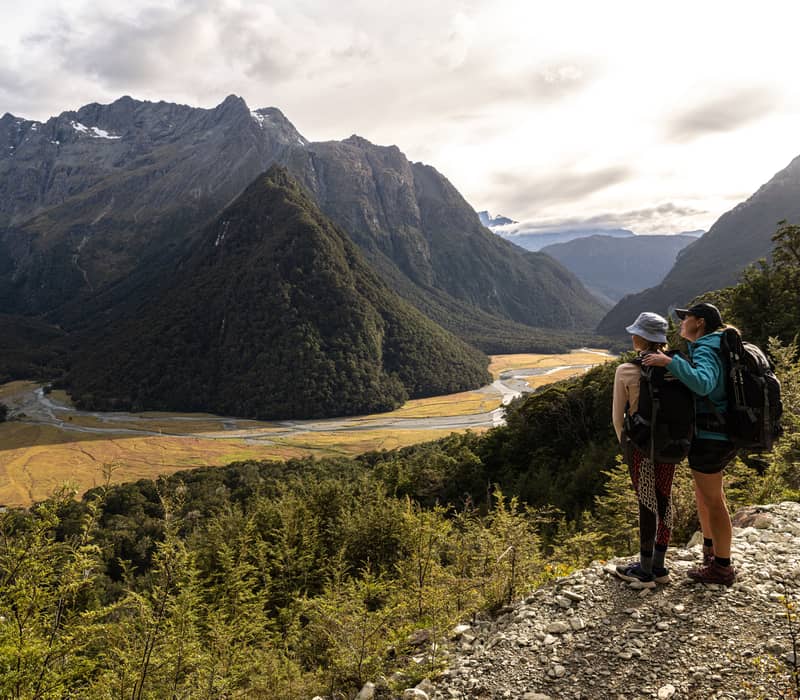 Two hikers with backpacks stand on a trail overlooking a river valley and tall mountains.