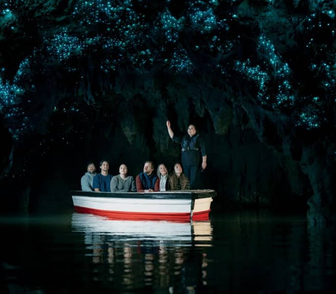 Group of people on a boat inside a dark cave illuminated by blue glowing lights of glowworms above.