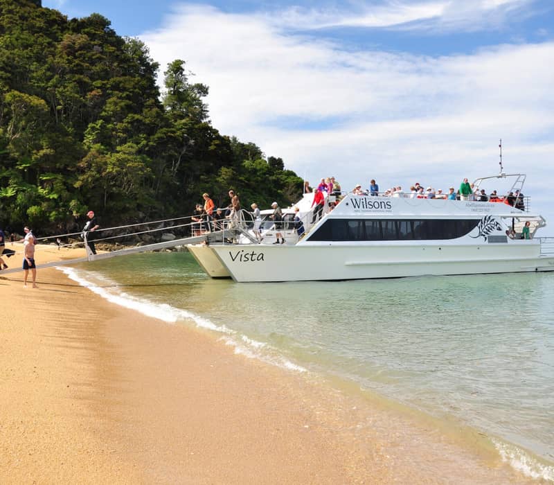 Tourists disembark from a white sightseeing boat onto a sandy beach with forested hills in Abel Tasman National Park.